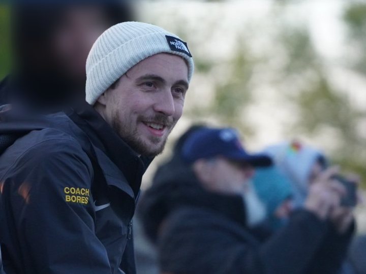 Ethan Bores at a meet, smiling toward the course, in a coach's jacket with his name on the sleeve.